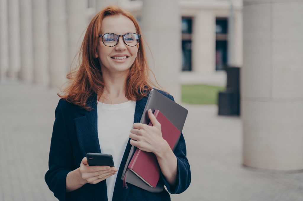 Happy red-haired business lady in official clothes standing outdoors, using mobile phone